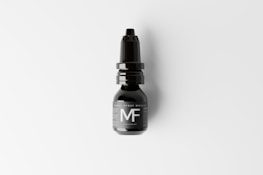 A black nasal spray bottle with a nozzle lid is placed against a light background. The label on the bottle features large, bold letters 'MF'. The design is sleek and minimalist, conveying a modern aesthetic.