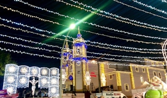 A church adorned with festive lights in various colors, including blue and gold. Strings of lights stretch across the night sky above, creating a vibrant and celebratory atmosphere. Illuminated decorations, like a depiction of Jesus on the cross, add to the festive scene.