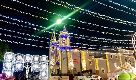 A church adorned with festive lights in various colors, including blue and gold. Strings of lights stretch across the night sky above, creating a vibrant and celebratory atmosphere. Illuminated decorations, like a depiction of Jesus on the cross, add to the festive scene.