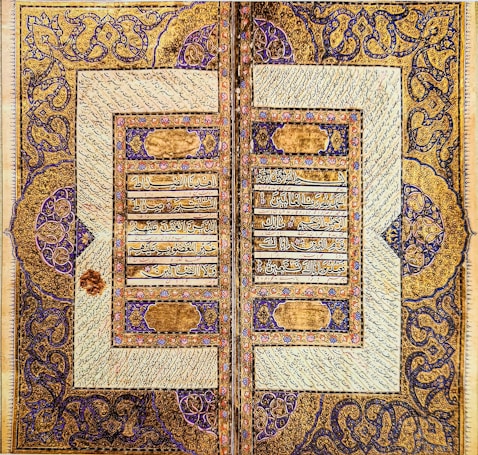 An intricately designed manuscript page featuring elaborate Arabic calligraphy bordered by symmetrical ornate patterns. The gold and blue patterns frame the text and are filled with floral motifs and geometric designs, displaying a high level of craftsmanship.