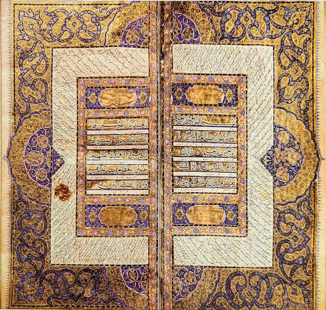 An intricately designed manuscript page featuring elaborate Arabic calligraphy bordered by symmetrical ornate patterns. The gold and blue patterns frame the text and are filled with floral motifs and geometric designs, displaying a high level of craftsmanship.