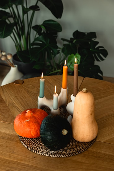 A wooden table features a woven placemat with three gourds: one orange, one dark green, and a long beige gourd. Four lit candles of varying heights and colors&mdash;blue, orange, and brown&mdash;are positioned on a modern, sculptural candle holder. In the background, there are lush green plants in a stylish vase, adding to the cozy and natural setting.