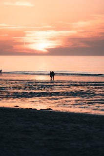 A serene beach scene at sunset with two people walking along the shore. The sky is filled with warm hues of orange and pink as the sun sets over the horizon. The calm sea reflects the colors of the sky, creating a peaceful and romantic atmosphere.