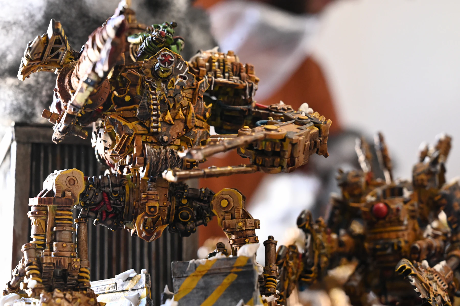 A painted walker model in yellow and rust, its body covered in pipes, plates and rivets