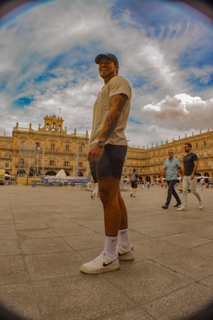 A person wearing casual sports attire, including a cap, is standing in a historic plaza with ornate architecture in the background. The sky is partly cloudy and the setting has a vibrant atmosphere with people walking around.