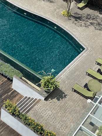A swimming pool with clear, calm water surrounded by a stone-tiled deck. To the side, there are two green lounge chairs and a small round table. Plants with yellow flowers line a section of the walkway, and a potted plant adds greenery near the pool.
