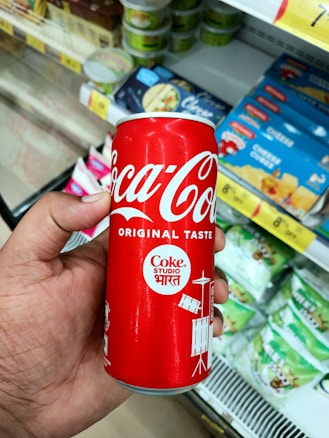 A hand holding a red Coca-Cola can with the logo and 'Original Taste' written on it. The background shows various packaged products on supermarket shelves.