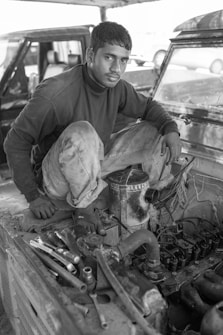 A person is sitting on the edge of a car engine bay, possibly working on the engine. The setting seems to be a workshop or garage. The individual is dressed in casual work clothes, which are slightly dirty, indicating hands-on work. Various tools and engine parts are visible around the person.