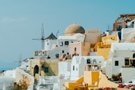 A picturesque view of a Greek island town featuring traditional Cycladic architecture. The scene includes whitewashed buildings with blue accents, dome-shaped roofs, and several yellow structures. A windmill is visible in the background along with people walking along the narrow streets. The sky is clear and bright, contributing to a vibrant and inviting atmosphere.