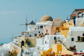 A picturesque view of a Greek island town featuring traditional Cycladic architecture. The scene includes whitewashed buildings with blue accents, dome-shaped roofs, and several yellow structures. A windmill is visible in the background along with people walking along the narrow streets. The sky is clear and bright, contributing to a vibrant and inviting atmosphere.