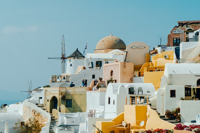 A picturesque view of a Greek island town featuring traditional Cycladic architecture. The scene includes whitewashed buildings with blue accents, dome-shaped roofs, and several yellow structures. A windmill is visible in the background along with people walking along the narrow streets. The sky is clear and bright, contributing to a vibrant and inviting atmosphere.