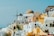 A picturesque view of a Greek island town featuring traditional Cycladic architecture. The scene includes whitewashed buildings with blue accents, dome-shaped roofs, and several yellow structures. A windmill is visible in the background along with people walking along the narrow streets. The sky is clear and bright, contributing to a vibrant and inviting atmosphere.