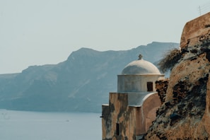 A historic stone building with a white dome overlooks the calm blue sea, with rugged cliffs and mountainous terrain in the background. The structure's aged walls have a rustic charm, enhanced by the natural landscape surrounding it.