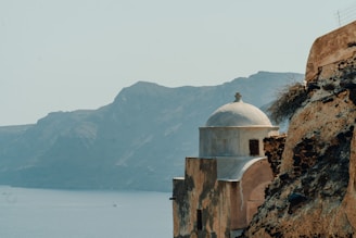 A historic stone building with a white dome overlooks the calm blue sea, with rugged cliffs and mountainous terrain in the background. The structure's aged walls have a rustic charm, enhanced by the natural landscape surrounding it.