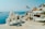 A picturesque coastal town features whitewashed buildings with domed roofs, set against a backdrop of a calm blue sea. A Greek flag waves gently in the breeze, and a church with distinctive bell towers stands prominently. Sunlight casts stark shadows across the structures, enhancing their architectural details.