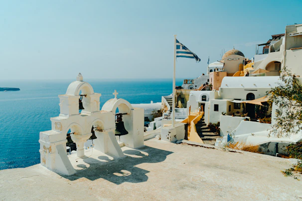 A picturesque coastal town features whitewashed buildings with domed roofs, set against a backdrop of a calm blue sea. A Greek flag waves gently in the breeze, and a church with distinctive bell towers stands prominently. Sunlight casts stark shadows across the structures, enhancing their architectural details.