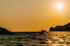 A skipper steering the boat while the sun sets, casting golden hues over the sea and silhouetting the coastline