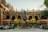 A historic courtyard with a central statue set in front of an ornate building featuring arched windows and rich stonework. Potted trees and several outdoor umbrellas provide shade for people seated at tables. Some patrons are enjoying their time at this atmospheric open-air café.