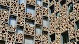 A building facade with a unique design featuring numerous round tubes of varying sizes arranged within rectangular frames. The tubes are predominantly tan and appear to be made of a material resembling bamboo or cardboard. The frames are set against a backdrop of silver or metallic panels.