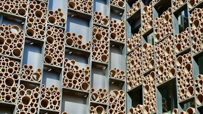 A building facade with a unique design featuring numerous round tubes of varying sizes arranged within rectangular frames. The tubes are predominantly tan and appear to be made of a material resembling bamboo or cardboard. The frames are set against a backdrop of silver or metallic panels.