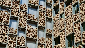 A building facade with a unique design featuring numerous round tubes of varying sizes arranged within rectangular frames. The tubes are predominantly tan and appear to be made of a material resembling bamboo or cardboard. The frames are set against a backdrop of silver or metallic panels.