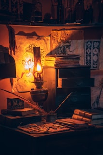 A dimly lit, vintage-style study desk illuminated by a glowing oil lamp casts warm, orange light on its surroundings. The desk is cluttered with an assortment of objects including old books, a quill, a pair of scissors, and some rolled parchment papers. Several artworks depicting mystical creatures hang on the wall above the desk. The overall ambiance is mysterious and old-fashioned.