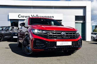 A bright red Volkswagen SUV is prominently displayed in front of a dealership with a sign reading 'Crewe Volkswagen.' The car has sleek black rims and a modern grill design, parked on a paved surface. Other vehicles are showcased in the background, contributing to a professional automotive setting.