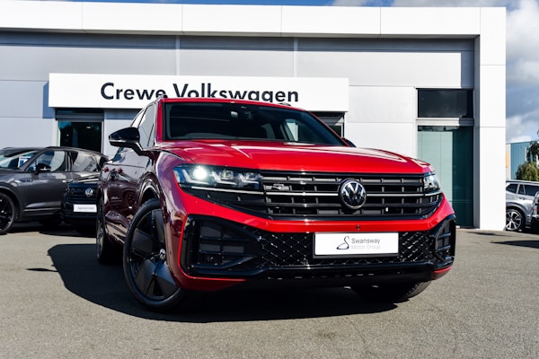 A bright red Volkswagen SUV is prominently displayed in front of a dealership with a sign reading 'Crewe Volkswagen.' The car has sleek black rims and a modern grill design, parked on a paved surface. Other vehicles are showcased in the background, contributing to a professional automotive setting.