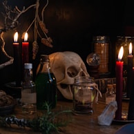 A dimly lit setting features a collection of objects associated with dark, mystical themes. Candles of red and black provide a flickering glow, creating a mysterious ambiance. A human skull serves as a macabre centerpiece, surrounded by jars filled with various items. Dried herbs hang above, while a crystal sphere and small potion bottles add to the enigmatic atmosphere.