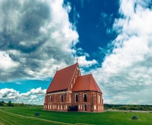 A red-bricked church with a large, sloped reddish-brown roof stands alone on a well-manicured green lawn. The structure features arched windows and crosses on its roof peaks. The sky above is vast and dramatic, filled with large, dense clouds with patches of blue sky visible.