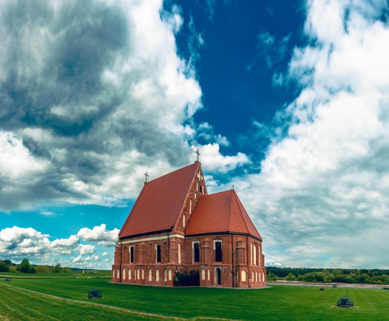 A red-bricked church with a large, sloped reddish-brown roof stands alone on a well-manicured green lawn. The structure features arched windows and crosses on its roof peaks. The sky above is vast and dramatic, filled with large, dense clouds with patches of blue sky visible.