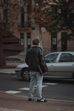 A person stands on the sidewalk wearing a black jacket, light blue jeans, and black sneakers with white stripes, holding a black folder. A silver car is passing by on the road. The background includes buildings, trees, and a large modern sculpture.