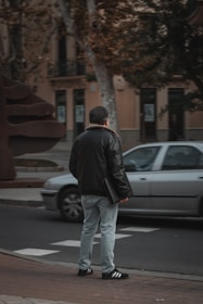 A person stands on the sidewalk wearing a black jacket, light blue jeans, and black sneakers with white stripes, holding a black folder. A silver car is passing by on the road. The background includes buildings, trees, and a large modern sculpture.