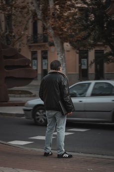 A person stands on the sidewalk wearing a black jacket, light blue jeans, and black sneakers with white stripes, holding a black folder. A silver car is passing by on the road. The background includes buildings, trees, and a large modern sculpture.