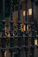 An ornate wrought iron fence with twisting bars is seen in front of a softly lit building. The background features rectangles of light, likely from windows or light fixtures, partially obscured by the grille. There is a hint of greenery behind the fence, suggesting plants or foliage.