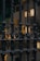 An ornate wrought iron fence with twisting bars is seen in front of a softly lit building. The background features rectangles of light, likely from windows or light fixtures, partially obscured by the grille. There is a hint of greenery behind the fence, suggesting plants or foliage.