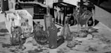A collection of clear and intricately designed glass bottles and vases displayed on a table. Around the glassware, there are also cardboard boxes containing sets of glassware, possibly champagne flutes. The setting is outdoors, possibly at a market or a display area.