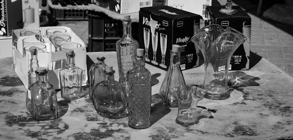 A collection of clear and intricately designed glass bottles and vases displayed on a table. Around the glassware, there are also cardboard boxes containing sets of glassware, possibly champagne flutes. The setting is outdoors, possibly at a market or a display area.