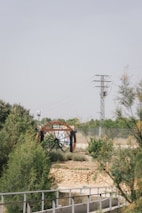 A large water wheel with metal beams set within a natural landscape featuring greenery and shrubs. A stone wall and metal railing are in the foreground, while an electricity pylon and wires stand in the background against a clear sky.