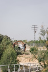 A large water wheel with metal beams set within a natural landscape featuring greenery and shrubs. A stone wall and metal railing are in the foreground, while an electricity pylon and wires stand in the background against a clear sky.