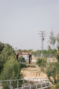 A large water wheel with metal beams set within a natural landscape featuring greenery and shrubs. A stone wall and metal railing are in the foreground, while an electricity pylon and wires stand in the background against a clear sky.