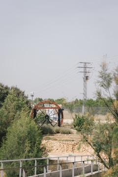 A large water wheel with metal beams set within a natural landscape featuring greenery and shrubs. A stone wall and metal railing are in the foreground, while an electricity pylon and wires stand in the background against a clear sky.
