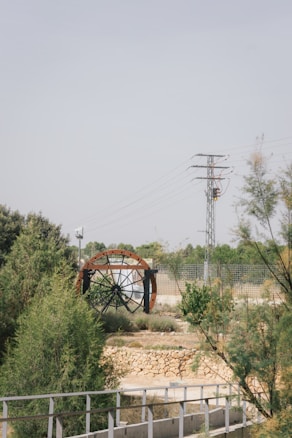 A large water wheel with metal beams set within a natural landscape featuring greenery and shrubs. A stone wall and metal railing are in the foreground, while an electricity pylon and wires stand in the background against a clear sky.