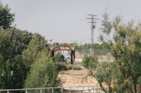 A wooden water wheel is surrounded by lush greenery and shrubs, positioned in an open area. In the background, there is a metal lattice electricity pylon with wires stretching across the sky. The setting appears rural and serene, with a dry stone wall visible in the vicinity.