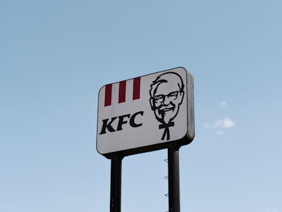 A tall roadside sign displays the KFC logo, featuring the company's name and a graphical portrait, set against a clear blue sky.