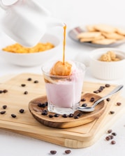 A stream of coffee is being poured from a white pot over a scoop of ice cream in a glass filled with a pink creamy liquid. Coffee beans are scattered on a wooden board, accompanied by a spoon. In the background, there are bowls containing biscuits and caramel popcorn, creating a pleasant dessert setting.