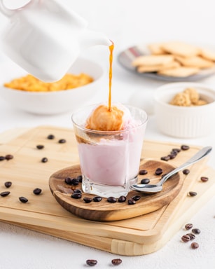 A stream of coffee is being poured from a white pot over a scoop of ice cream in a glass filled with a pink creamy liquid. Coffee beans are scattered on a wooden board, accompanied by a spoon. In the background, there are bowls containing biscuits and caramel popcorn, creating a pleasant dessert setting.