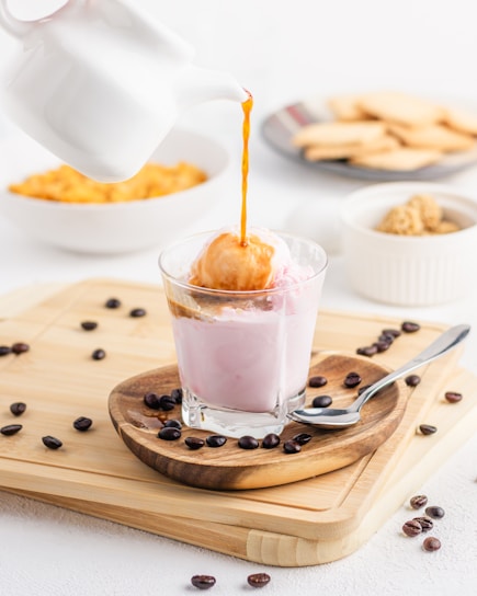 A stream of coffee is being poured from a white pot over a scoop of ice cream in a glass filled with a pink creamy liquid. Coffee beans are scattered on a wooden board, accompanied by a spoon. In the background, there are bowls containing biscuits and caramel popcorn, creating a pleasant dessert setting.