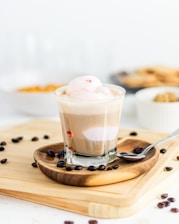 A glass cup filled with a layered coffee drink topped with a scoop of ice cream. It is placed on a wooden coaster with scattered coffee beans around the base. In the background, there are blurred elements including a bowl of crackers and another bowl partially visible, suggesting a casual dining setting.