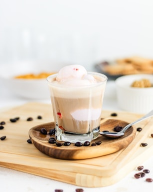 A glass cup filled with a layered coffee drink topped with a scoop of ice cream. It is placed on a wooden coaster with scattered coffee beans around the base. In the background, there are blurred elements including a bowl of crackers and another bowl partially visible, suggesting a casual dining setting.
